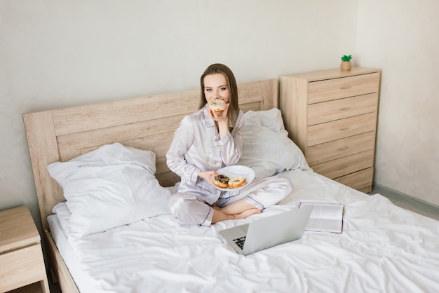 Young woman in white towel and robe in kitchen during quarantine. Hold donut on plate. Breakfast time at home.