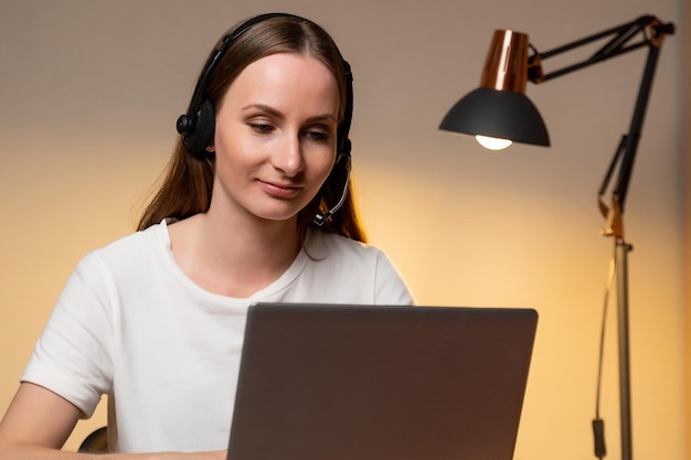 Young woman in a white Tshirt and headset is working on a laptop computer at home Work at home Video conference Video Call Online class for students