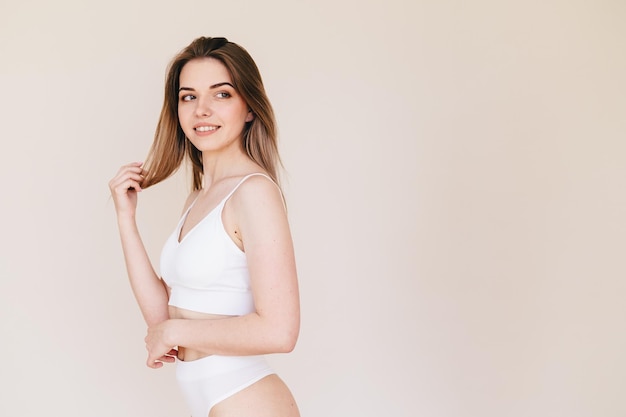 Young Woman in White Underwear Stands Sideways on a Beige Background and Cares for Her Hair