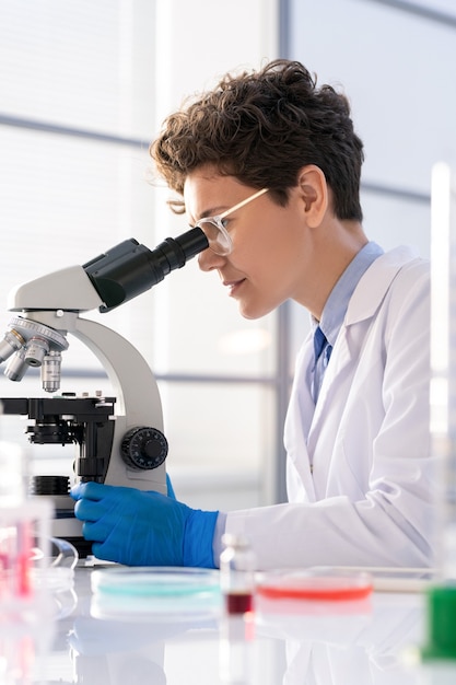 Young woman in whitecoat and gloves looking in microscope while studying charcteristics of new chemical substances in laboratory