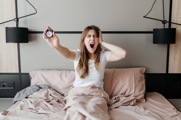 Young woman with alarm clock in hand waking up in the morning in bed at home