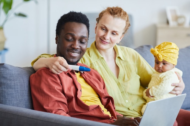 Young woman with baby on her knees showing pregnancy test to her husband while they sitting at home