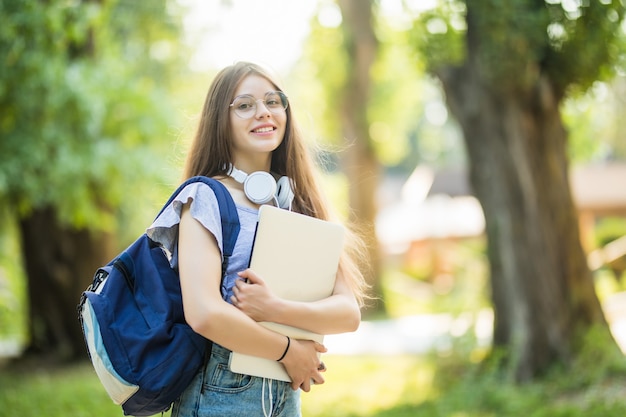 Young woman with backpack walking through park with silver laptop in hands