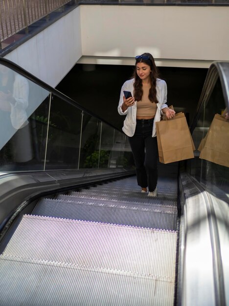 Young woman with bags and mobile phone going up the escalator to the shopping centre