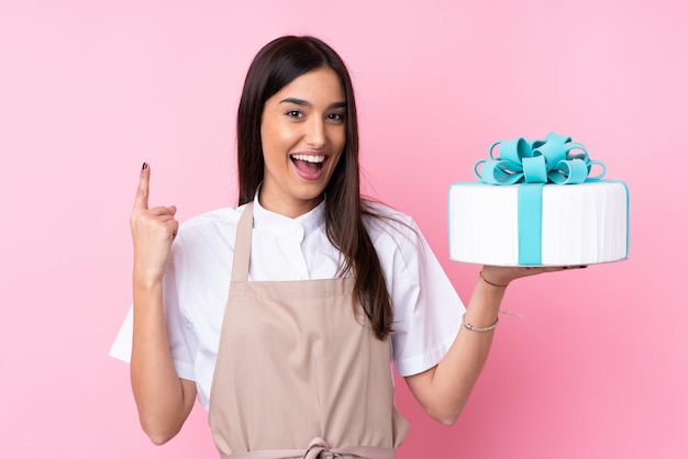 Young woman with a big cake over isolated wall pointing up a great idea