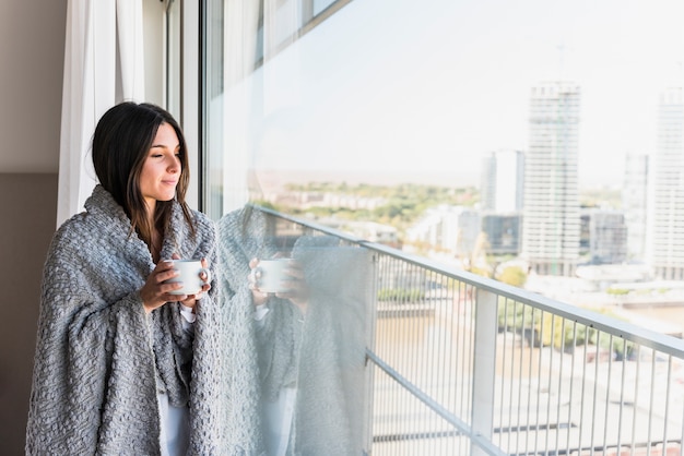 Young woman with blanket over her shoulder holding coffee cup in hand looking away