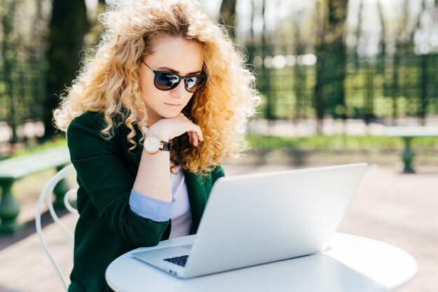 Young woman with blond curly hair wearing sunglasses and elegant green jacket sitting in front of open laptop outdoors reading news online having pensive expression People technology concept