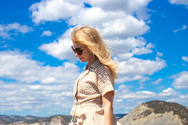 A young woman with blond hair in sunglasses bowed her head against a blue sky with clouds in a mountainous area