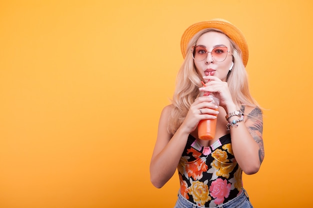 Young woman with blonde hair drinking orange juice with sunglasses in studio over yellow background