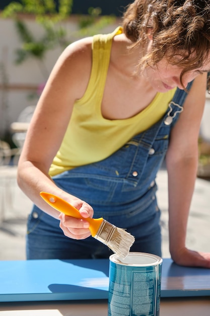 Young woman with blue denim overalls and yellow tshirt in the garden dipping a brush in paint to paint a piece of furniture