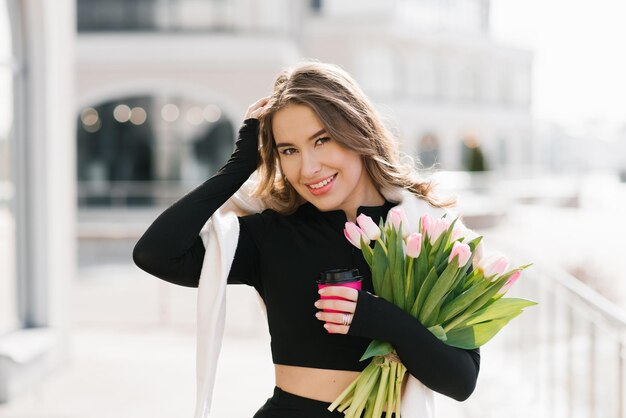 Young woman with a bouquet of spring tulips and a cup of coffee walks down the street in the city