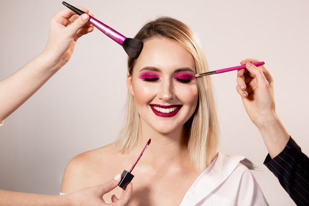 Young woman with bright dark pink makeup and eyes closed posing in studio background. Makeup artist's hands correct makeup with a special brush. Professional makeup