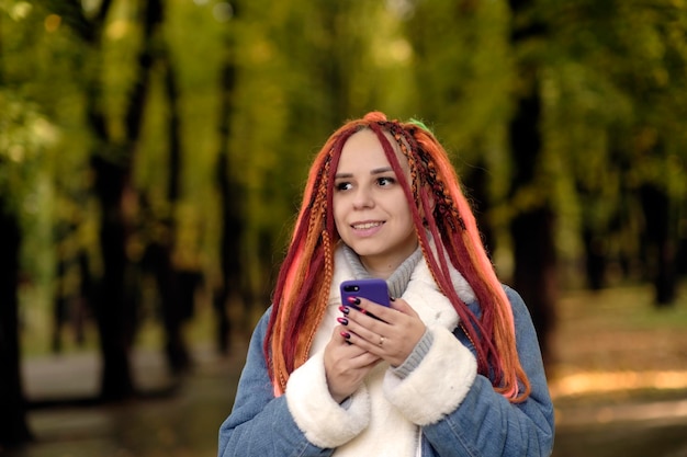 Young woman with bright dreadlocks browsing mobile phone standing in forest park Vivid female with multicolored hairstyle using smartphone on walk