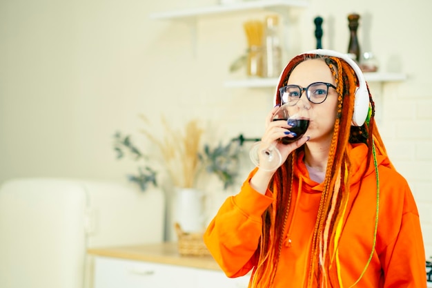 Young woman with bright dreadlocks in glasses wireless headphones with glass of red wine in kitchen