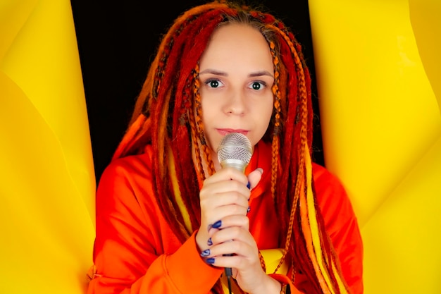 Young woman with bright dreadlocks singing into microphone sticking out of hole of on yellow background Emotional female singer with mic in studio