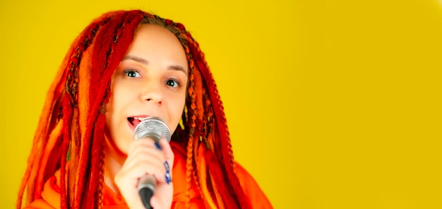 Young woman with bright dreadlocks singing into microphone on yellow background Emotional female singer with mic in studio Close up