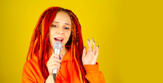 Young woman with bright dreadlocks singing into microphone on yellow background Emotional female singer with mic in studio Close up