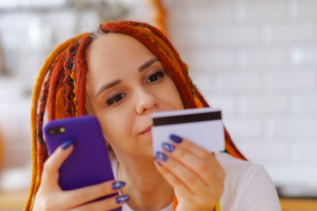 Young woman with bright dreadlocks with mobile phone credit card sitting in kitchen Happy female browsing smartphone entering card details paying internet purchase Concept of online shopping