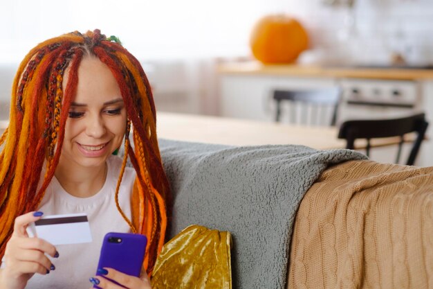 Young woman with bright dreadlocks with mobile phone credit card sitting in living room Happy female browsing smartphone entering card details paying internet purchase Concept of online shopping