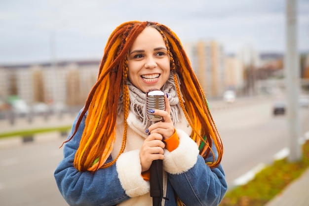 Young woman with bright dreadlocks with retro microphone on city street Portrait of female singer singing into microphone in cloudy weather