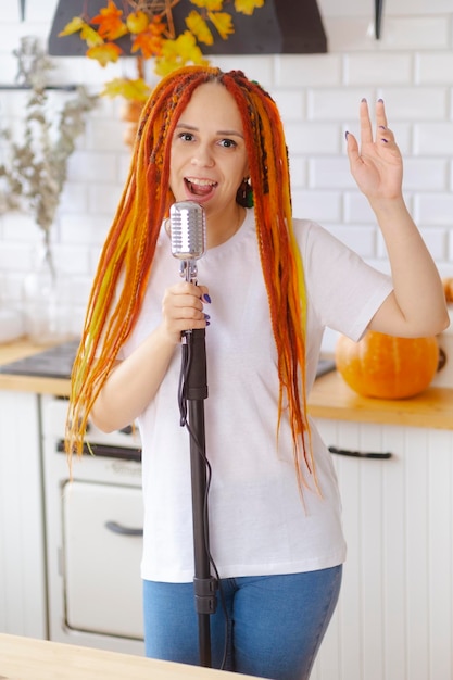 Young woman with bright hairstyle with retro microphone in kitchen Portrait of female singer with dreadlocks singing into microphone at home