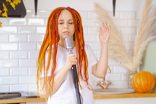 Young woman with bright hairstyle with retro microphone in kitchen Portrait of female singer with dreadlocks singing into microphone at home