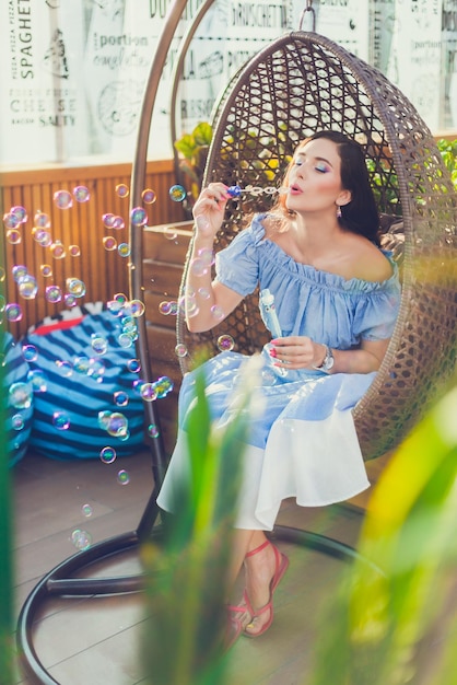 A young woman with bright makeup is sitting in a summer outdoor cafe in a hanging chair and blowing soap bubbles