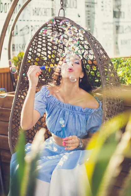 A young woman with bright makeup is sitting in a summer outdoor cafe in a hanging chair and blowing soap bubbles