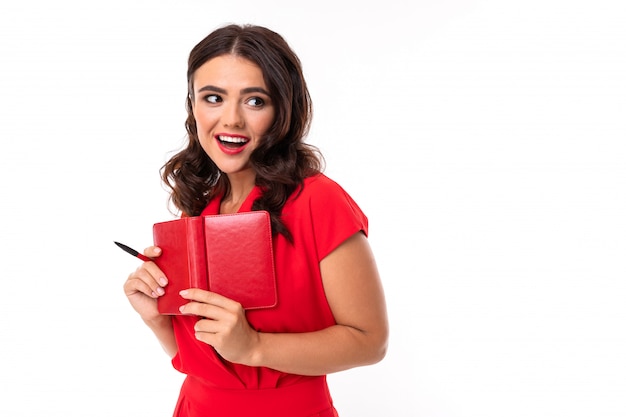 A young woman with bright makeup, in a red summer dress stands with a notebook and smiles