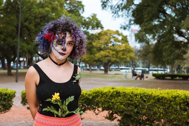 Young woman with catrina makeup and folkloric clothes stands in the park looking at the camera