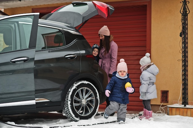 Young woman with children hold eco bags and charging electric car with open trunk in the yard of her house