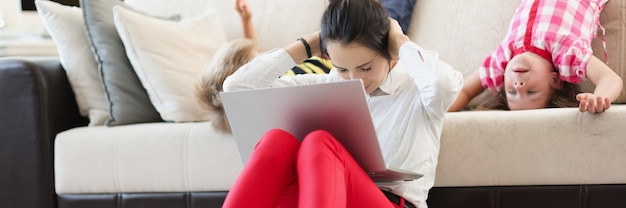 Young woman with children sitting on floor with laptop on her lap and covering her ears