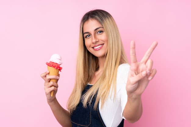 Young woman with a cornet ice cream over isolated pink wall smiling and showing victory sign