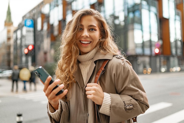 Young woman with curly blonde hair using the phone with a cup of coffee in hands on the city streets