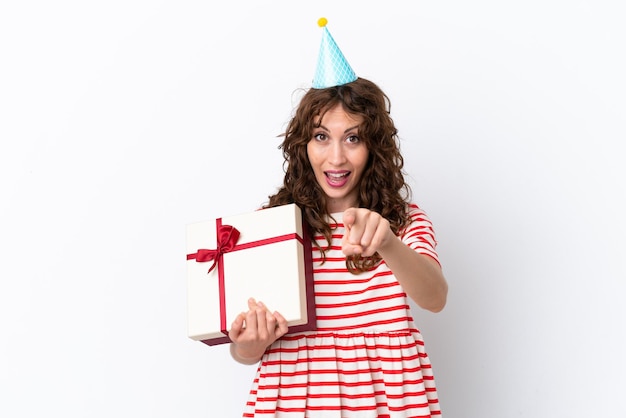 Young woman with curly hair holding present isolated on white background surprised and pointing front