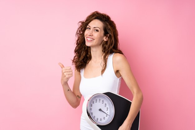 Young woman with curly hair holding a weighing machine over isolated pink wall