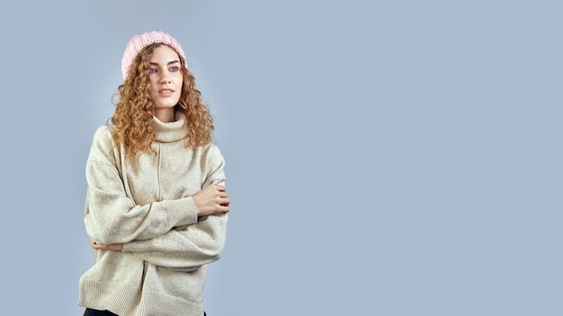 A young woman with curly hair poses in a hat and sweater