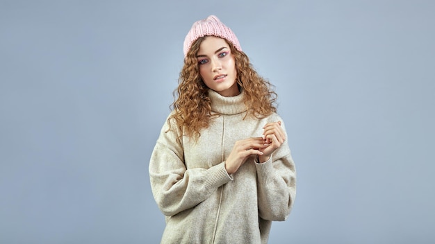 A young woman with curly hair poses in a hat and sweater