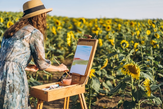 A young woman with curly hair and wearing a hat is painting in nature. A woman stands in a sunflower field on a beautiful day