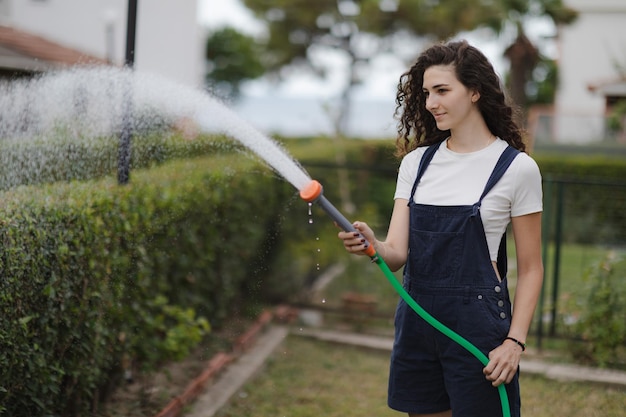 Young woman with curly hair wearing overall watering plants in country house backyard garden care ecofriendly lifestyle suburban life concept