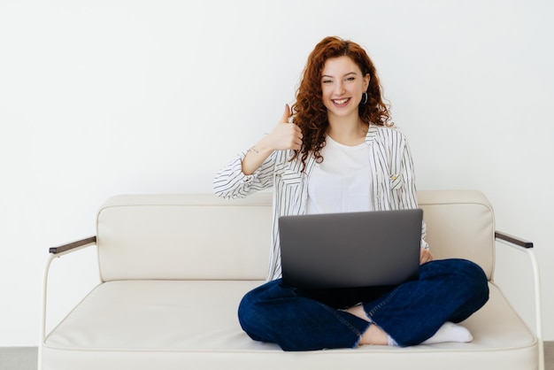 Young woman with curly red hair is lying on the couch and participating in a video conference on a laptop freelance and remote work