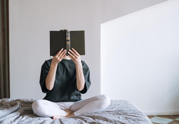 Young woman with dark long hair in casual clothes reading book on bed at the home