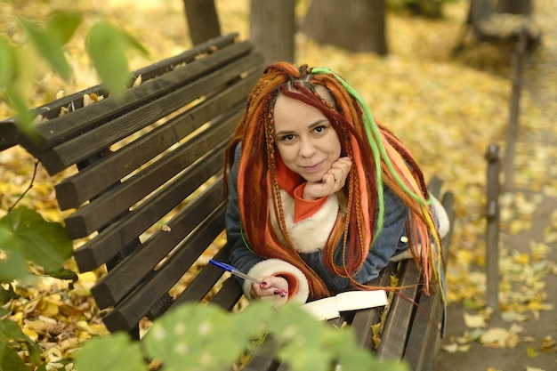 Young woman with dreadlocks drawing writing in notepad lying on park bench in golden autumn