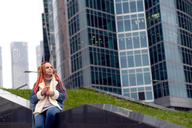 Young woman with dreadlocks drinking coffee on background of skyscrapers Relaxed female hipster enjoying hot drink sitting on stairway in downtown