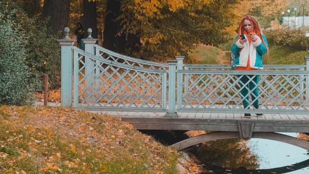 Young woman with dreadlocks drinking coffee from cup browsing mobile phone standing on wooden bridge over creek in park in golden autumn