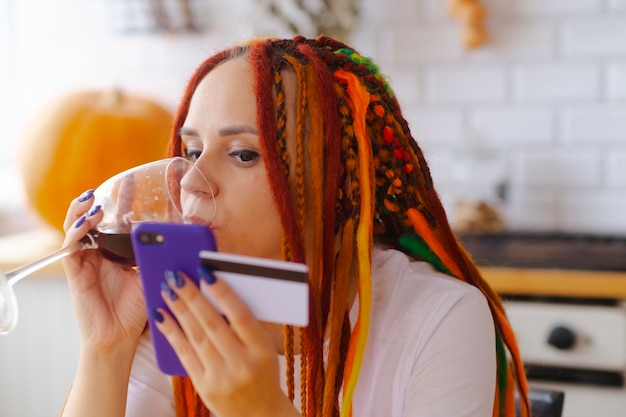 Young woman with dreadlocks drinking red wine from glass browsing mobile phone entering card details paying internet purchase sitting in kitchen