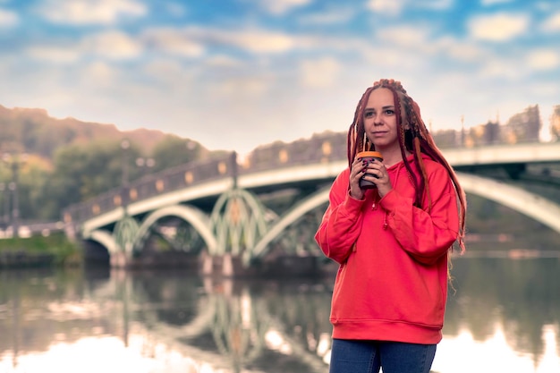 Young woman with dreadlocks holding cup of coffee standing near river lake on background of big bridge in city park