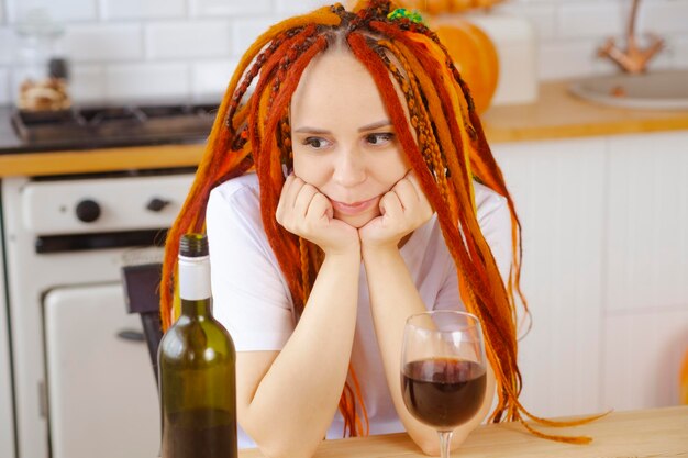 Young woman with dreadlocks looking at wine propping face in hands sitting in kitchen Adult female drinking red wine from glass relaxing at home