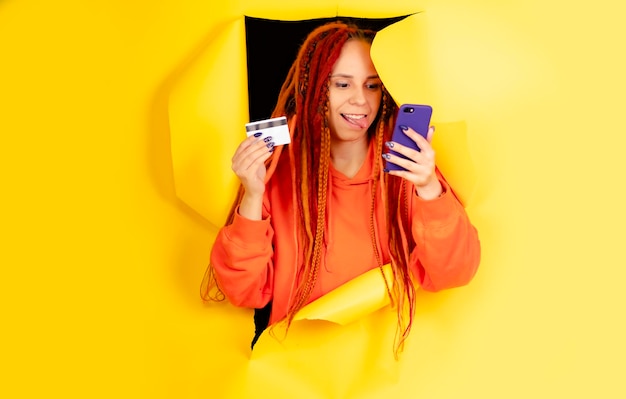 Young woman with dreadlocks mobile phone credit card sticking out of hole of on yellow background Positive female browsing smartphone entering card details paying internet purchase