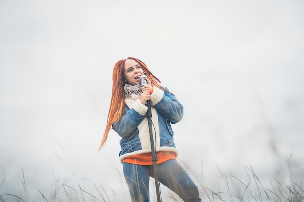 Young woman with dreadlocks singing into microphone on background of cloudy sky Female singer enjoying singing in nature